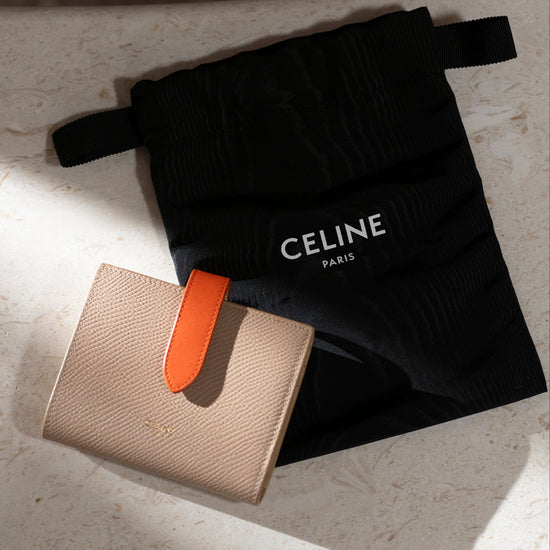 Beige Celine compact wallet with orange accent and black Celine pouch on a light surface