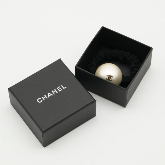 A Chanel-branded black box with a removable lid, containing a hair scrunchie adorned with a faux pearl with a 'CC' Chanel logo.