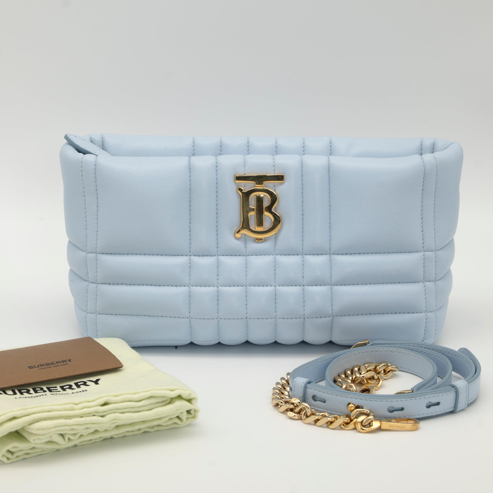 Light blue quilted Burberry handbag with gold 'TB' logo, chain strap, and Burberry dust bag on a white background
