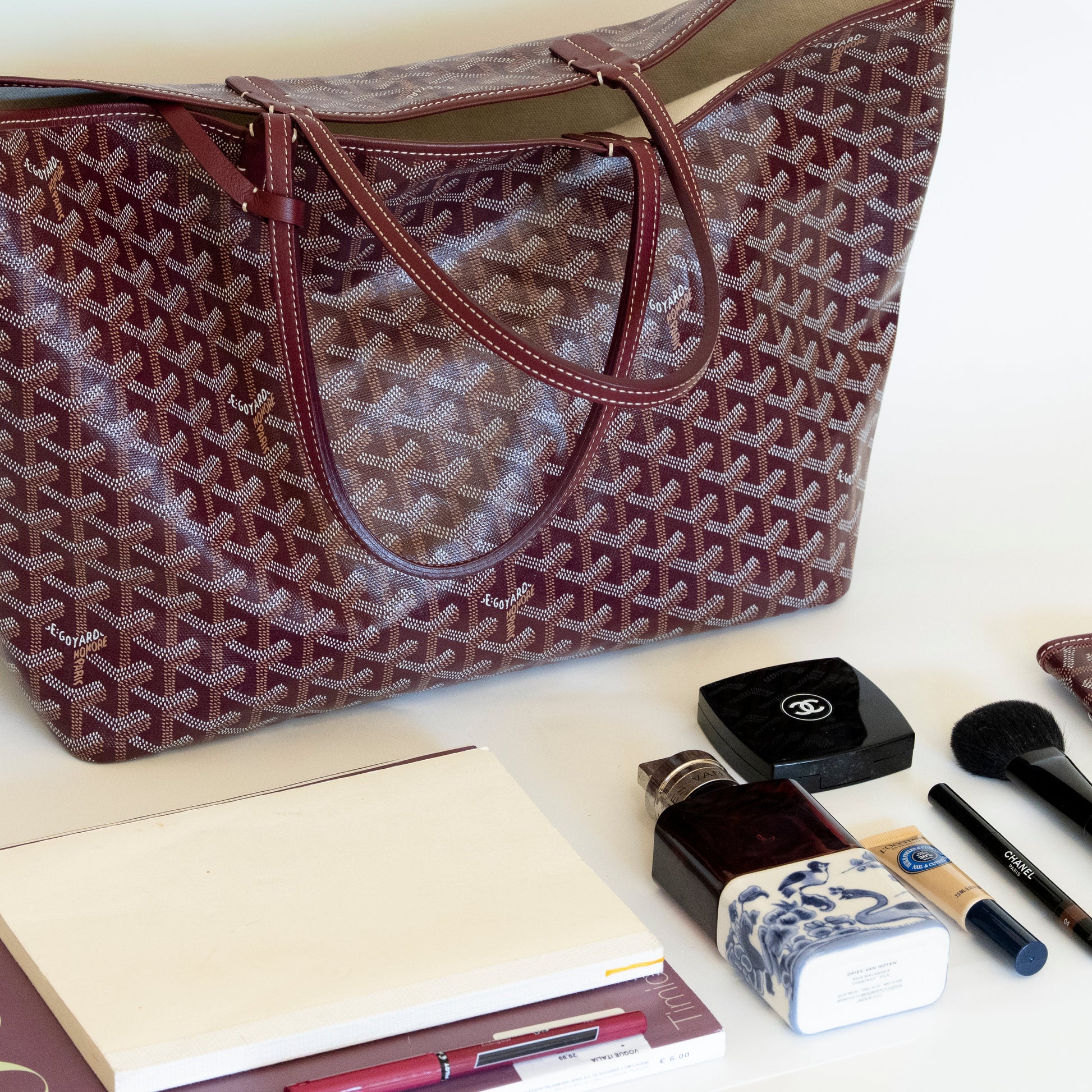 Burgundy Goyard tote with makeup items on a white surface