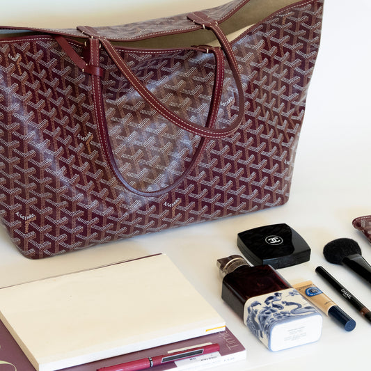 Burgundy Goyard tote with makeup items on a white surface